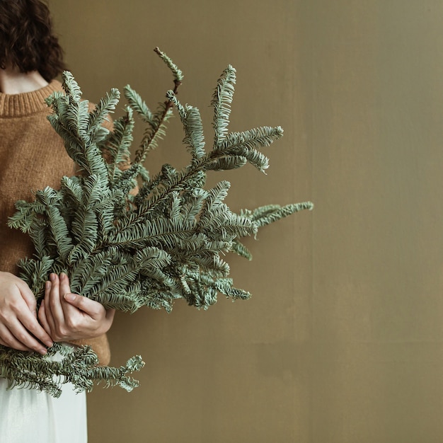 Mujer hermosa joven en suéter y falda sosteniendo ramas de abeto contra la pared de olivo. Concepto de Navidad festiva de moda minimalista.