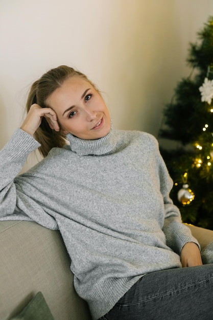 Mujer hermosa joven en suéter gris de punto sentado en el sofá cerca del árbol de Navidad.