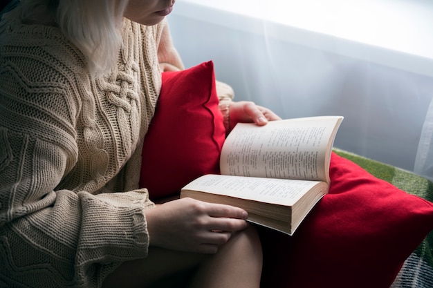 Mujer hermosa joven en un suéter de punto y medias leyendo un libro sentado en el sofá