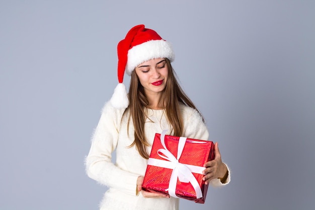 Mujer hermosa joven en suéter con sombrero de navidad sosteniendo regalos sobre fondo gris en estudio