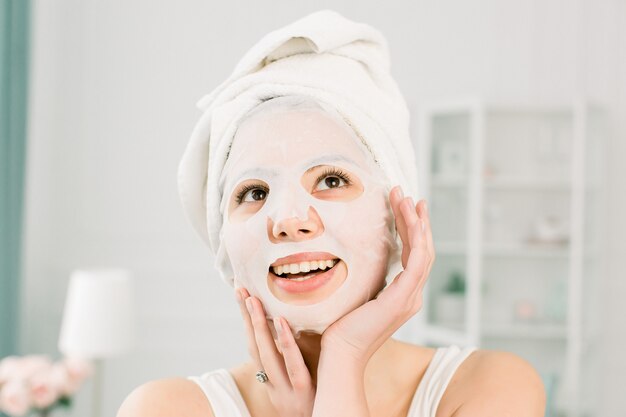 Mujer hermosa joven con la toalla blanca en la cabeza, aplicando en una mascarilla, concepto del cuidado de piel. Spa, cuidado de la salud. Mujer con máscara purificadora en su rostro