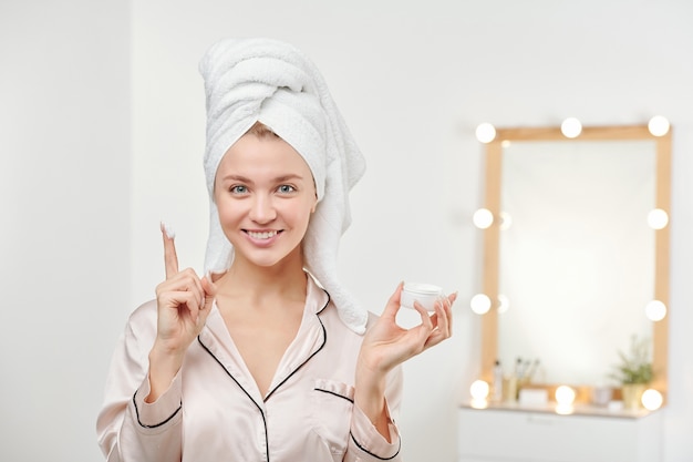 Foto mujer hermosa joven con toalla blanca en la cabeza va a aplicar crema hidratante en el rostro después de la ducha matutina