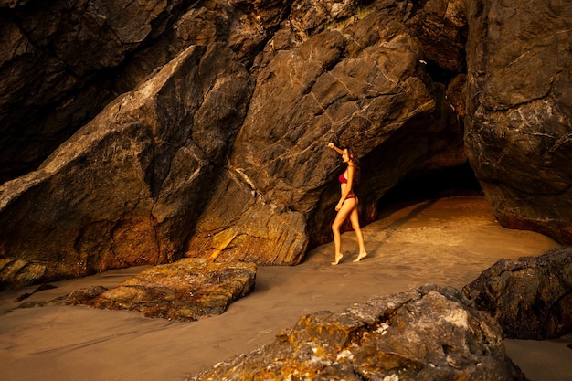 Mujer hermosa joven en traje de baño bikini rojo posando de pie sobre la arena, junto a las rocas en la playa en Goa India.