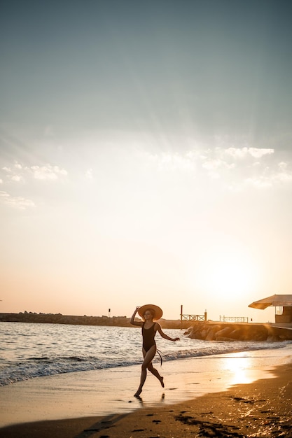 Mujer hermosa joven en traje de baño negro y sombrero con gafas camina por la playa al atardecer. El concepto de recreación en el mar. Enfoque selectivo