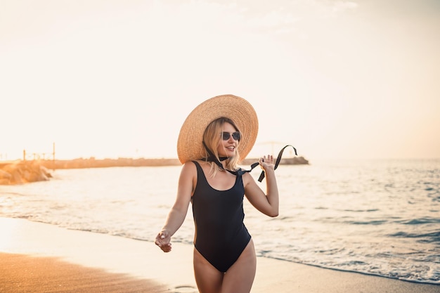 Mujer hermosa joven en traje de baño negro y sombrero con gafas camina por la playa al atardecer El concepto de recreación marina Enfoque selectivo