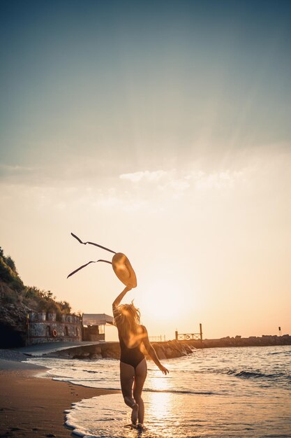 Mujer hermosa joven en traje de baño negro y sombrero con gafas camina por la playa al atardecer El concepto de recreación marina Enfoque selectivo