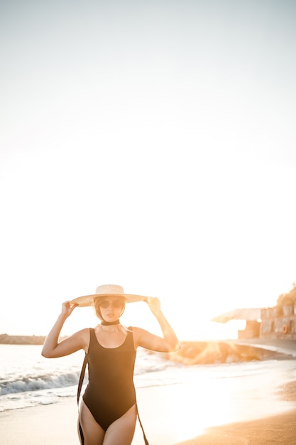 Mujer hermosa joven en traje de baño negro y sombrero con gafas camina por la playa en Turquía al atardecer. El concepto de recreación en el mar. Enfoque selectivo