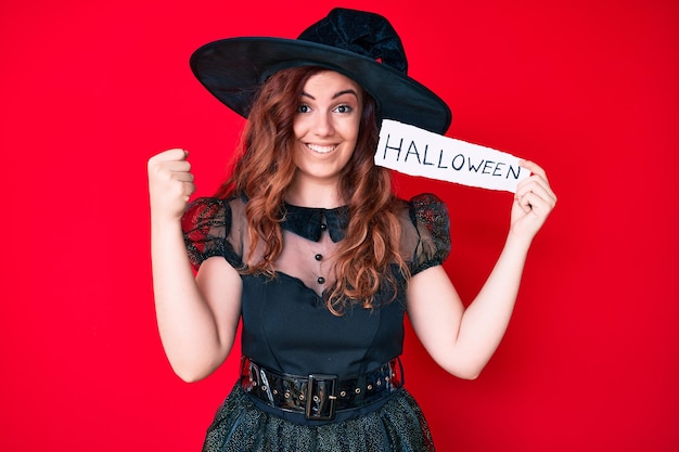 Mujer hermosa joven con traje de bruja sosteniendo papel con mensaje de halloween gritando orgullosa celebrando la victoria y el éxito muy emocionada con los brazos levantados