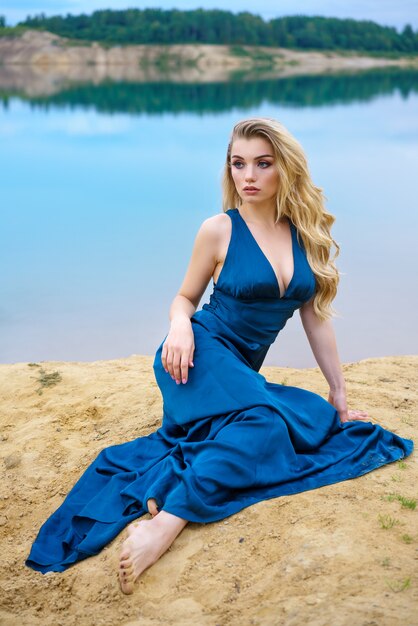 Una mujer hermosa joven triste con un vestido largo azul está sentada sola en la orilla del lago