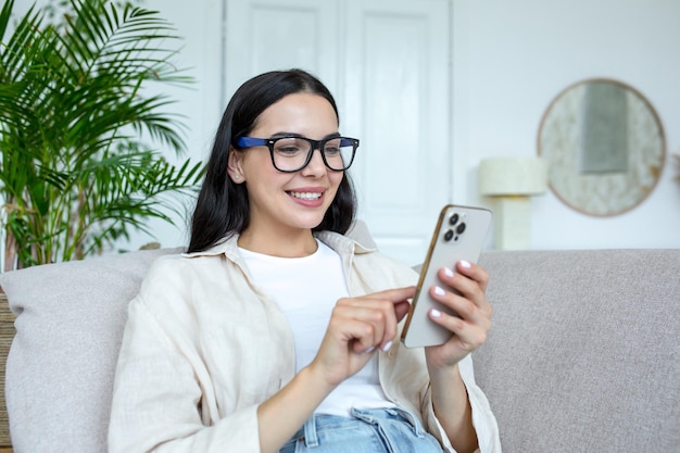 Mujer hermosa joven usando morena de teléfono en casa sentada en el sofá en la sala de estar y sonriendo feliz