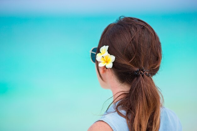 Mujer hermosa joven durante vacaciones en la playa tropical. Disfruta de unas vacaciones de verano solo en la playa con flores de frangipani en el pelo