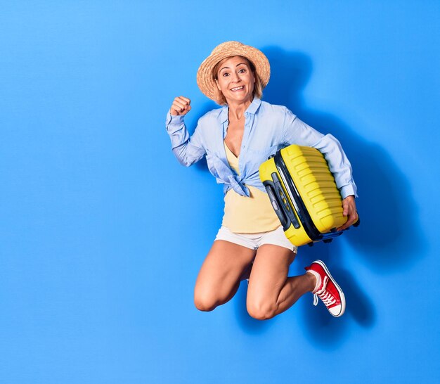 Mujer hermosa joven de vacaciones con ropa de verano y sombrero sonriendo feliz saltando con una sonrisa en la cara sosteniendo una bolsa de cabina sobre un fondo aislado