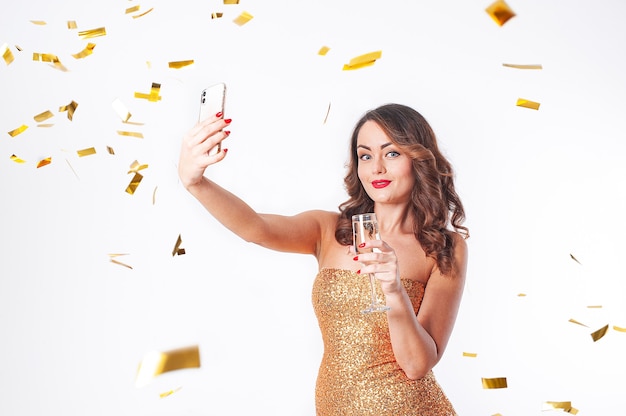 Mujer hermosa joven con un vestido dorado bebe champán, hace un selfie en un teléfono inteligente, divirtiéndose en una fiesta con confeti dorado sobre un fondo blanco