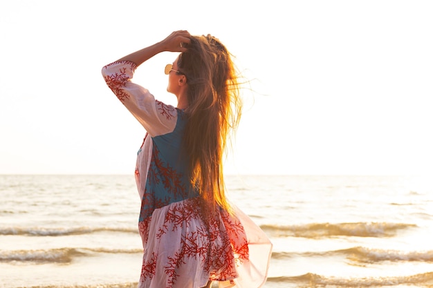 Mujer hermosa joven con vestido hermoso está caminando por la orilla del mar durante la puesta de sol