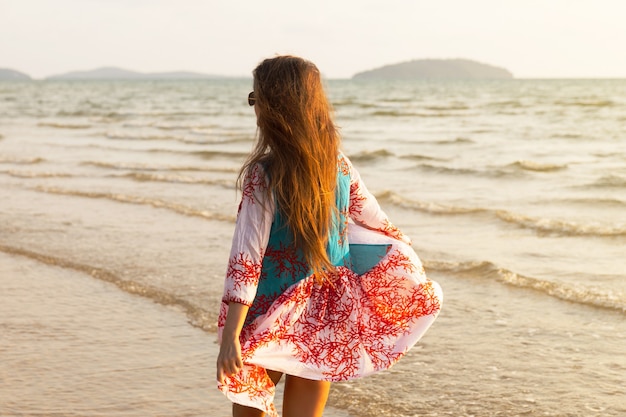 Mujer hermosa joven con vestido hermoso está caminando por la orilla del mar durante la puesta de sol