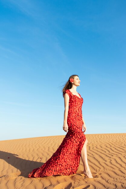 Mujer hermosa joven en vestido largo rojo con pétalos de rosas rojas entre el desierto Concepción de la rosa del desierto