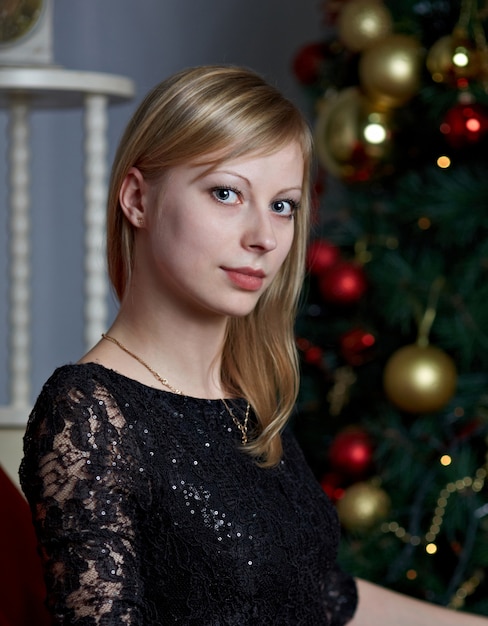 Mujer hermosa joven en vestido de noche elegante negro sentado en una silla cerca del árbol de Navidad en la víspera de año nuevo.