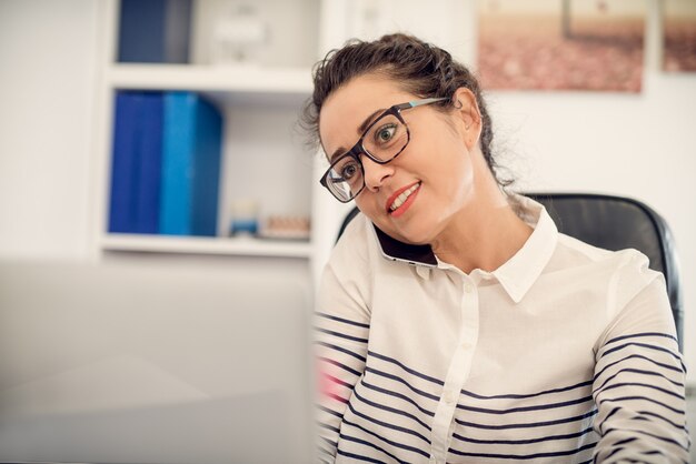 Mujer hermosa de mediana edad trabajadora hablando por un teléfono y mirando la computadora en su oficina.