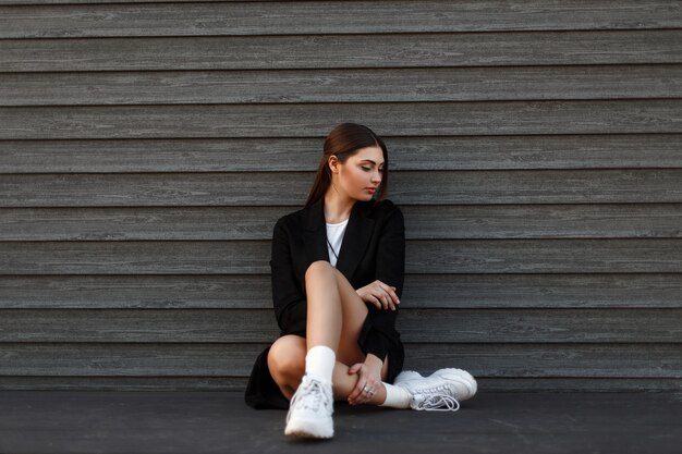 Mujer hermosa modelo en abrigo negro de moda con zapatillas blancas se sienta cerca de una pared de madera