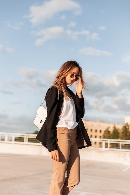 Mujer hermosa moderna en ropa de negocios de moda con bolsa camina en el estacionamiento y endereza gafas. Chica muy atractiva en elegante vestido casual en un día soleado en el cielo azul de fondo.