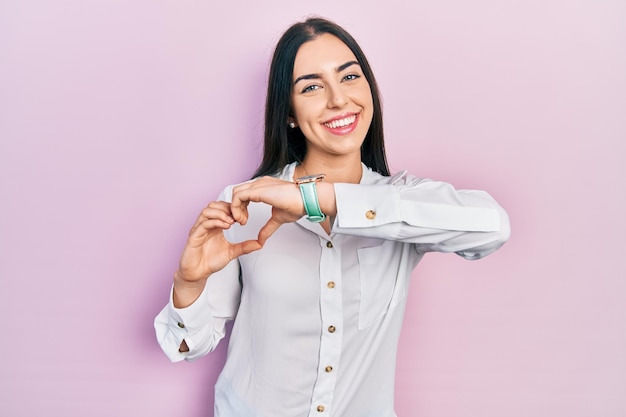 Mujer hermosa con ojos azules con camisa blanca casual sonriendo en el amor mostrando el símbolo del corazón y la forma con las manos concepto romántico