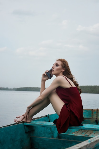 Mujer hermosa pelirroja de moda sentarse en el barco. Chica de retrato romántico de belleza en vestido rojo vinoso en barco de madera en el lago