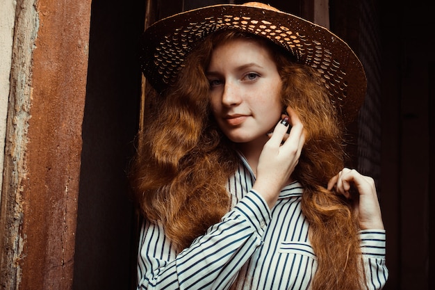 Mujer hermosa pelirroja con pelo largo y rizado en sombrero de paja posando cerca de la puerta de madera vieja