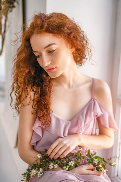 Mujer hermosa con el pelo rojo que se sienta en la ventana con las flores, humor de la primavera