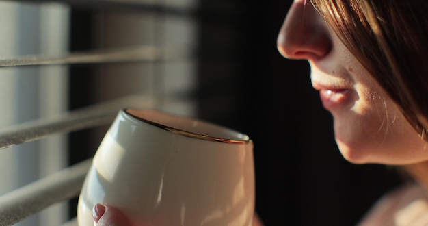 Mujer hermosa de pie cerca de la ventana con persianas por la mañana y bebiendo café o té