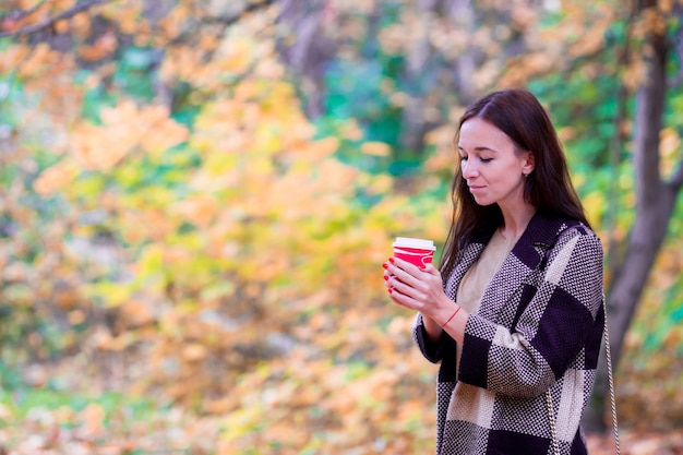 Mujer hermosa que bebe el café en parque del otoño debajo del follaje de otoño.