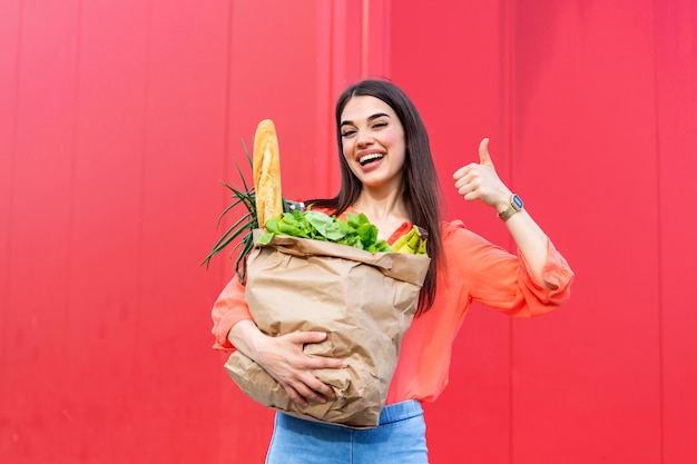 Mujer hermosa que sostiene bolsos de compras en el fondo rojo que muestra los pulgares para arriba.