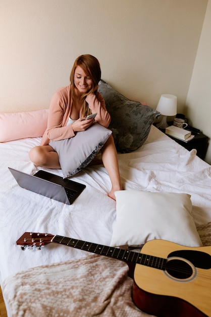 Mujer hermosa que usa el móvil y la computadora portátil en el dormitorio.