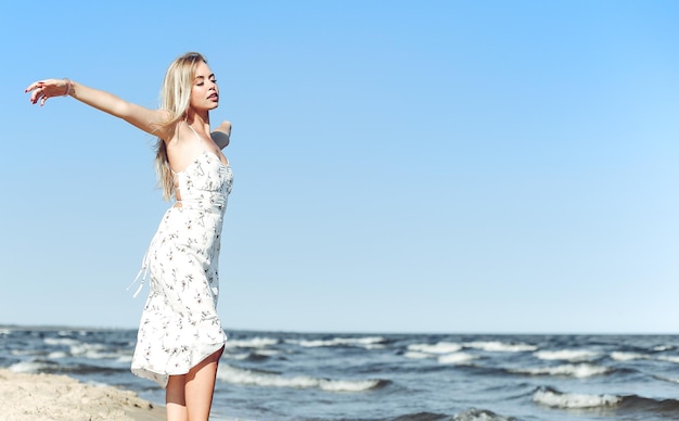 Mujer hermosa rubia feliz en la playa del océano de pie en un vestido blanco de verano, brazos abiertos.