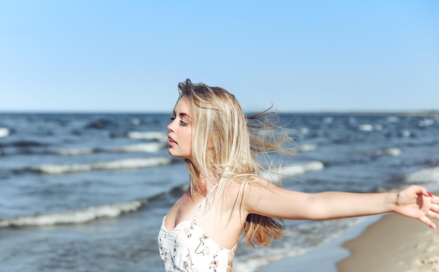 Mujer hermosa rubia feliz en la playa del océano de pie en un vestido blanco de verano, levantando las manos