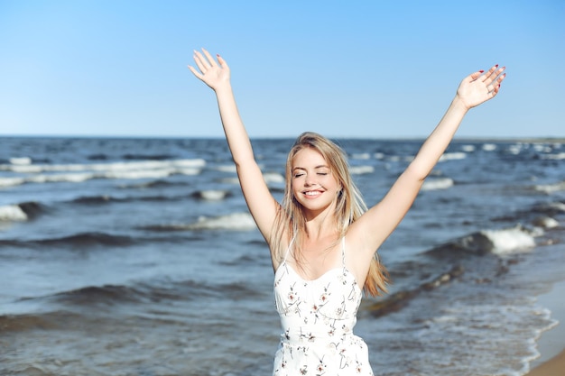 Mujer hermosa rubia feliz en la playa del océano de pie en un vestido blanco de verano, levantando las manos.