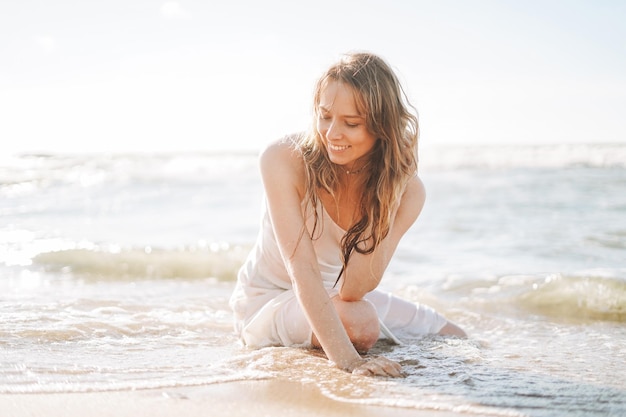 Mujer hermosa rubia joven con el pelo largo en vestido blanco disfrutando de la vida en la playa del mar