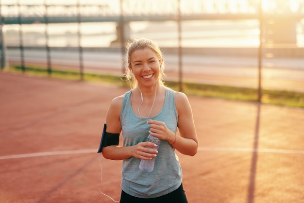 Mujer hermosa rubia sonriente con los oídos inn de los auriculares que sostienen la botella y que se colocan en la corte por la mañana.