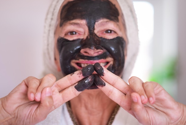 Mujer hermosa senior borrosa con una máscara de carbón facial de desintoxicación enfoque sonriente casero en los dedos cuidar el concepto de piel