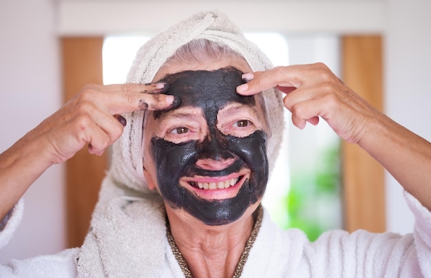 Mujer hermosa senior despreocupada aplicando una máscara de carbón facial de desintoxicación casera sonriendo cuidar el concepto de piel