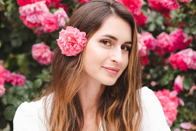 Mujer hermosa sonriente con cabello largo y rubio y flor de rosa sobre el fondo de la naturaleza en flor al aire libre