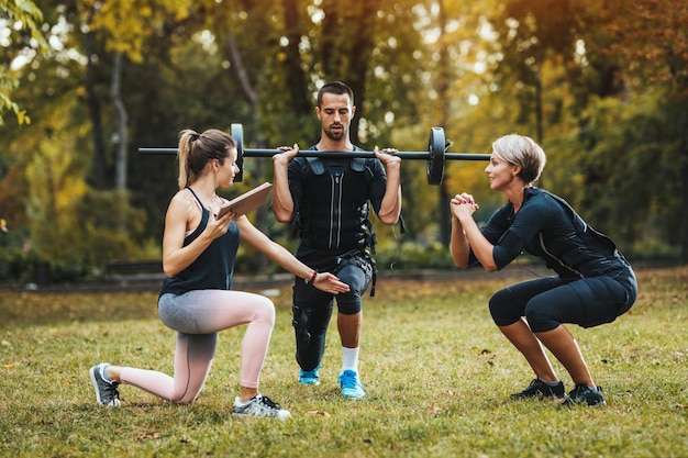 Una mujer hermosa y su pareja están haciendo ejercicios de piernas con un entrenador personal en el parque juntos, vestidos con un traje negro con un simulador electrónico EMS para estimular sus músculos.