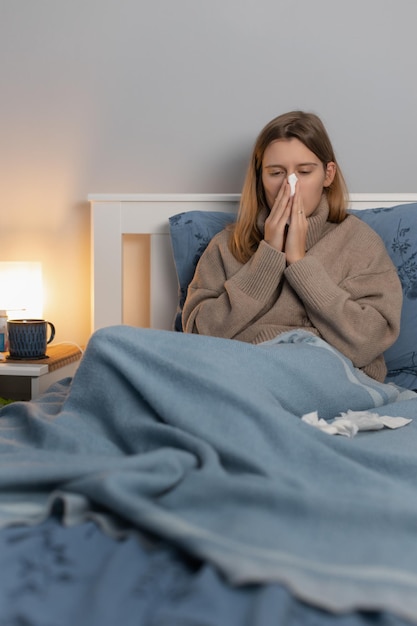 Mujer hermosa en suéter beige sentado en la cama y se suena la nariz en una servilleta de papel. Niña que sufre de secreción nasal, rinitis o sinusitis. Enfermedad estacional. Tratamiento a domicilio. Concepto de salud.