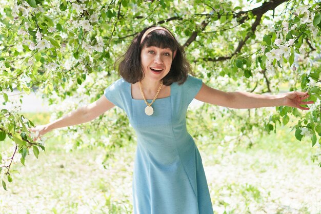 Una mujer hermosa con un vestido azul debajo de un árbol en flor en el jardín en primavera. La morena sonríe y está feliz