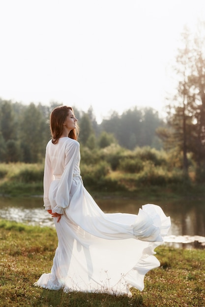 Mujer hermosa en un vestido blanco largo de pie cerca del lago