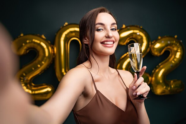 Mujer hermosa en vestido moderno de lujo haciendo selfie con champán frente a globos dorados isol ...