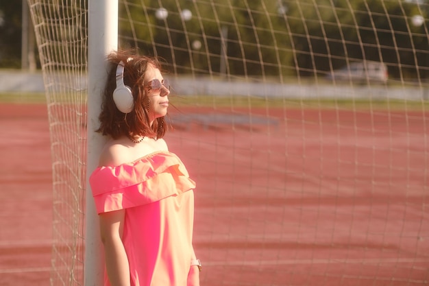 Una mujer hermosa con un vestido naranja y gafas de sol escucha música con auriculares blancos en el estadio apoyado en un gol de fútbol