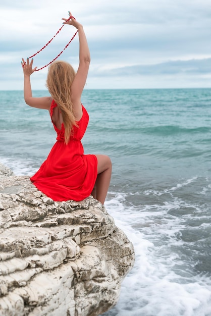 Una mujer hermosa con un vestido rojo sobre una roca blanca junto al mar en verano