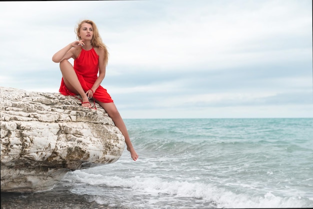 Una mujer hermosa con un vestido rojo sobre una roca blanca junto al mar en verano