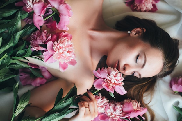 Foto una mujer hermosa yace sobre un paño blanco con peonías rosas en su cuerpo