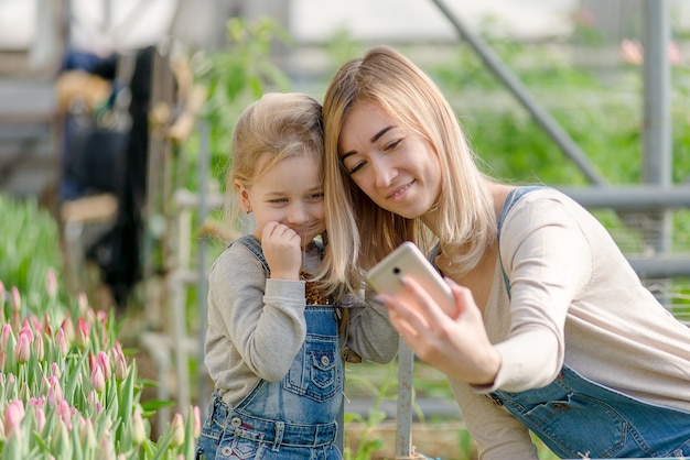 Una mujer con una hija pequeña se toma un selfie en un invernadero con flores en primavera.